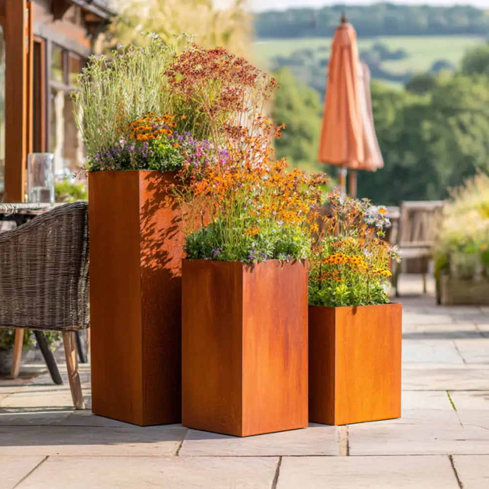 Three rust-coloured corten steel planters with flowers on a patio
