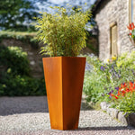Tall rust-colored planter with green plant in a garden setting
